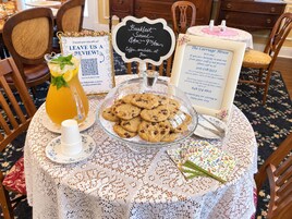 A table with a glass pitcher of iced tea, a bowl of cookies, a sign for breakfast service, and a welcome sign for The Carriage House.