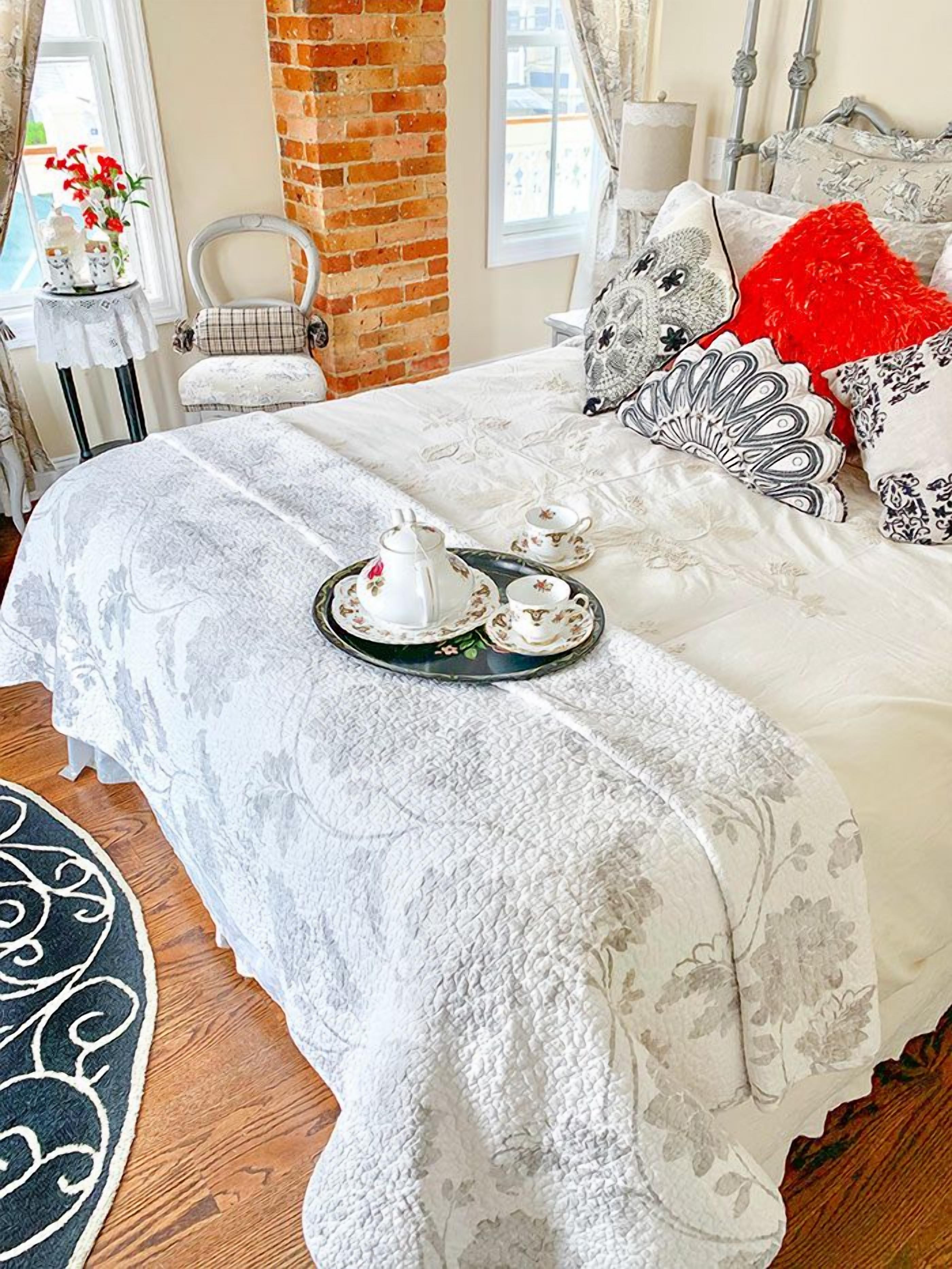 A neatly made bed with a tray of teacups and saucers, a chair, and a table with a vase of flowers.