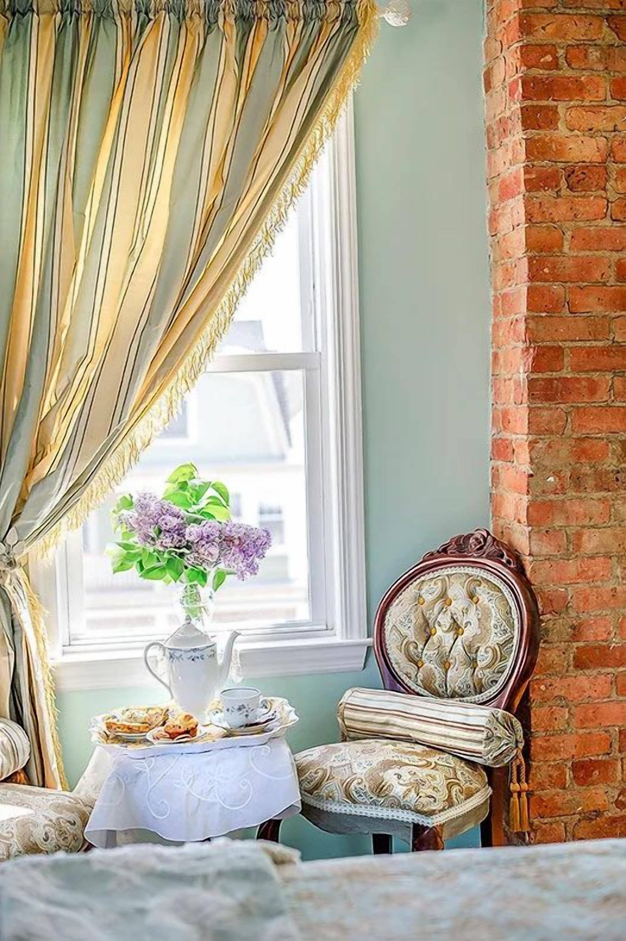 A room with a brick wall, a window with curtains, a table with a vase of flowers, and a chair with a patterned cushion.