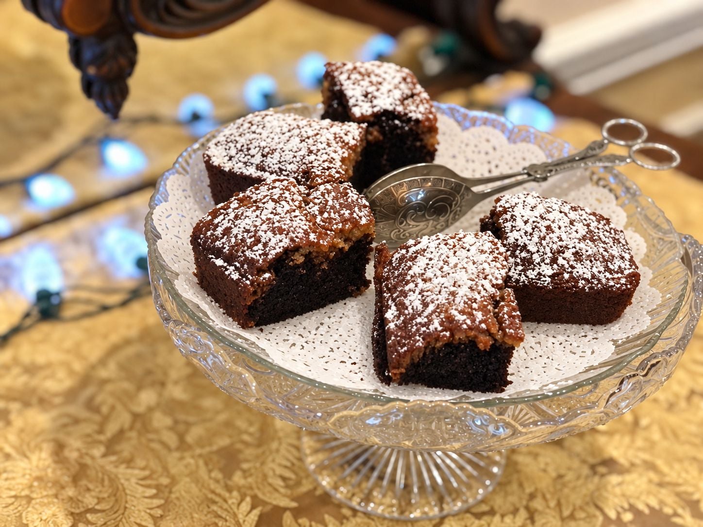 A glass cake stand with a lace doily, holding several square-shaped desserts dusted with powdered sugar.