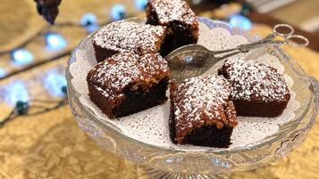 A glass cake stand with a lace doily, holding several square-shaped desserts dusted with powdered sugar.