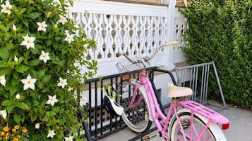 A pink bicycle parked on a porch with a white railing and a green plant beside it.