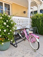 A pink bicycle parked on a porch with a white railing and a green plant beside it.