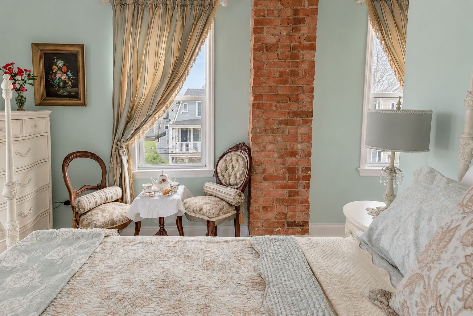 A bedroom with a brick wall, a white dresser, two chairs, a small table with a teapot, and a bed with a patterned bedspread.