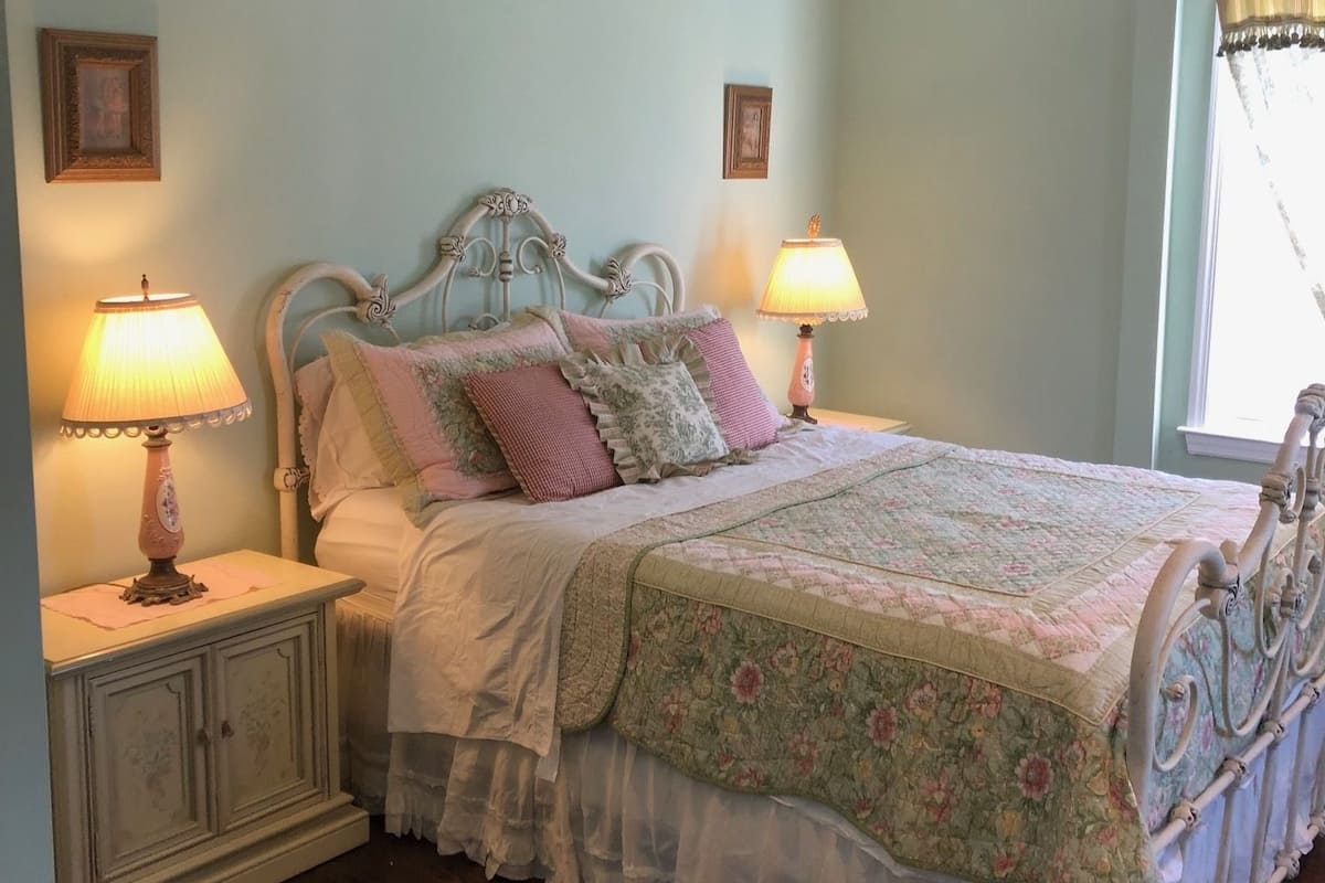 A bedroom with a bed, two bedside tables with lamps, a window with curtains, and a picture on the wall.