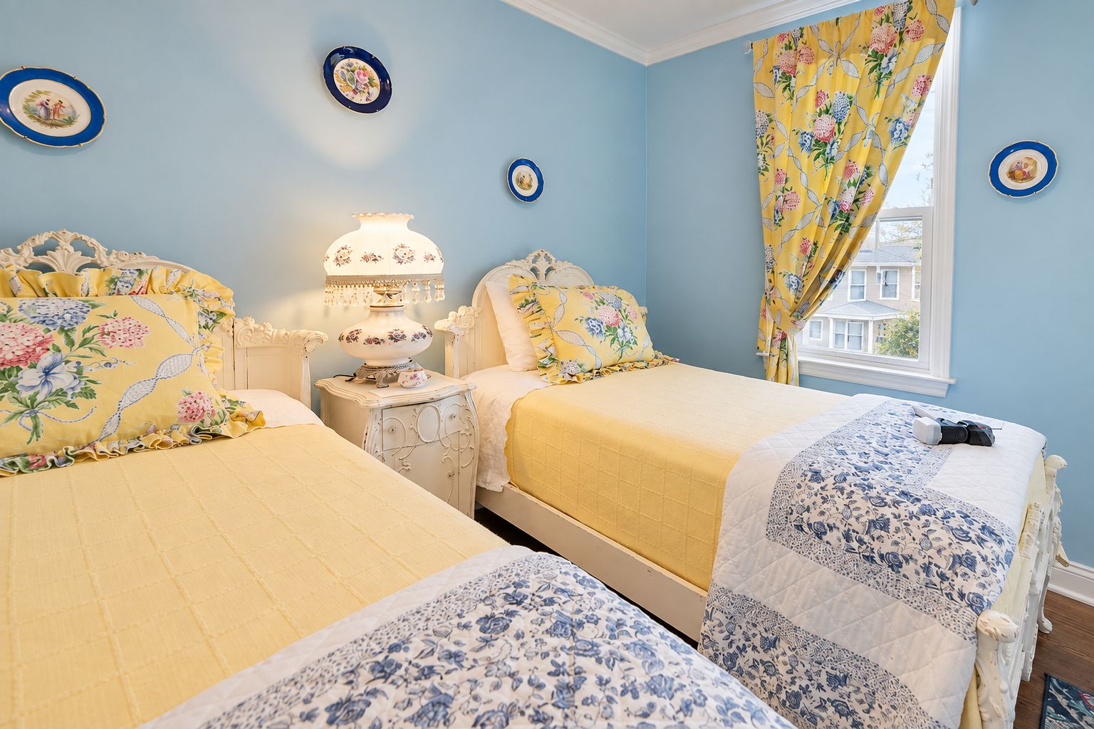 Un dormitorio con dos camas, una mesita de noche, una lámpara y cortinas con estampado floral.