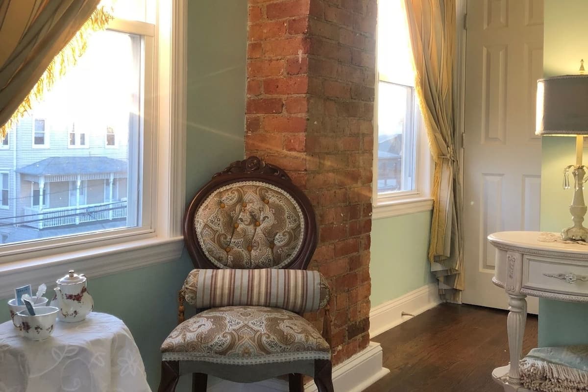 A room with a brick fireplace, a dining table with tea set, and a chair with a patterned cushion.