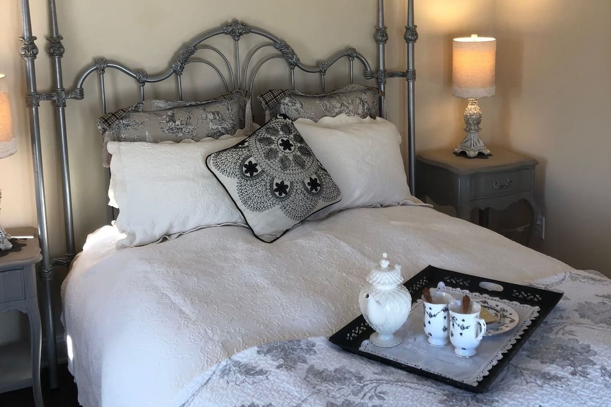 A neatly made bed with a decorative headboard, a tray with teapot and cups, and a nightstand with a lamp.