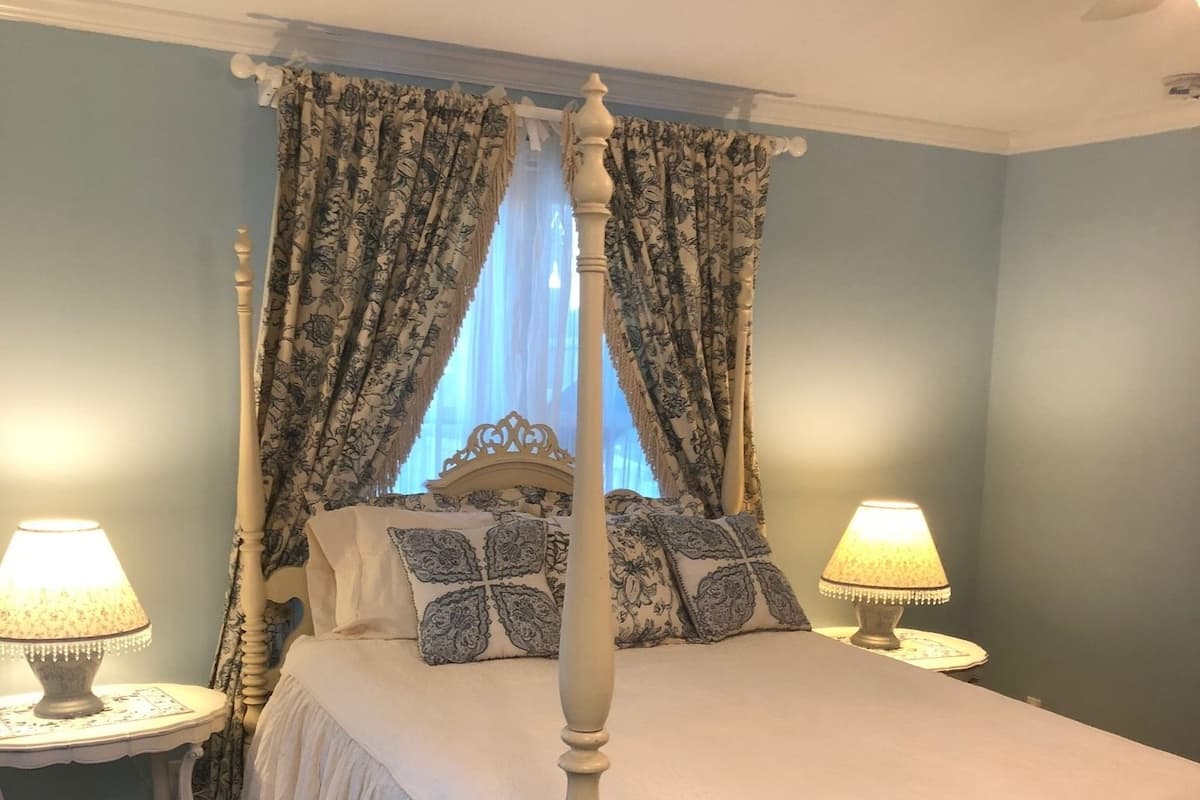 A four-poster bed with a canopy, two bedside tables with lamps, patterned curtains, and a window with sheer drapes.