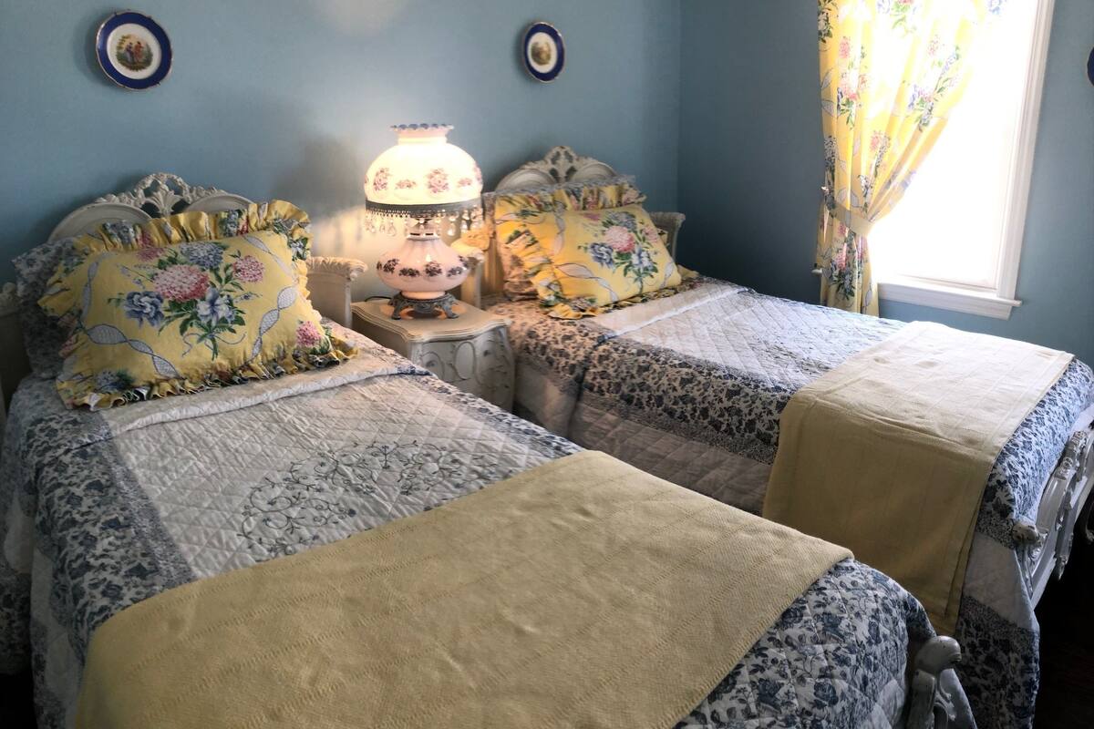 A bedroom with two beds, floral bedding, a bedside table with a lamp, and a window with curtains.