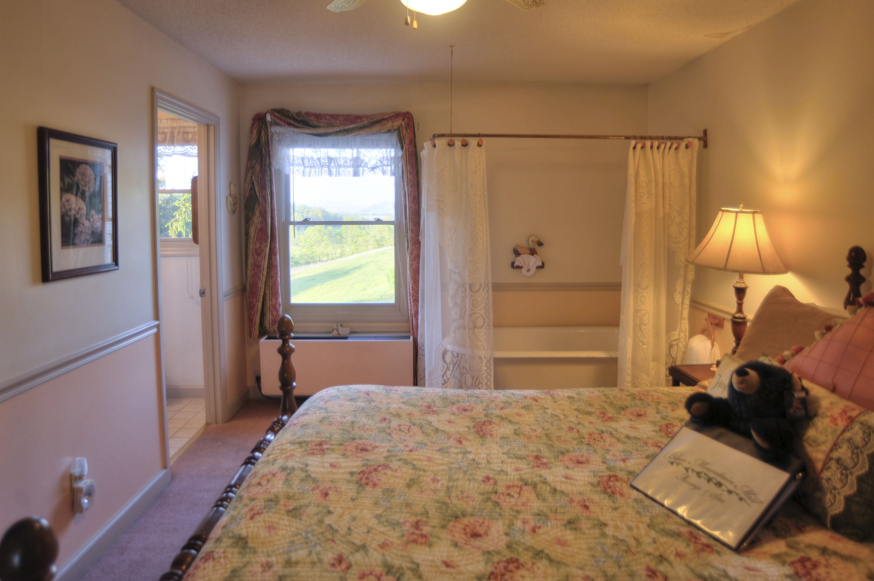 A bedroom with a floral bedspread, a teddy bear on the bed, a window with curtains, a bedside lamp, and a framed picture on the wall.