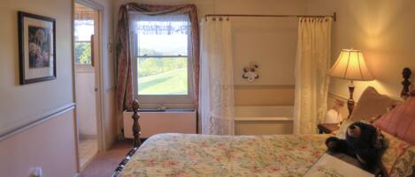 A bedroom with a floral bedspread, a teddy bear on the bed, a window with curtains, a bedside lamp, and a framed picture on the wall.