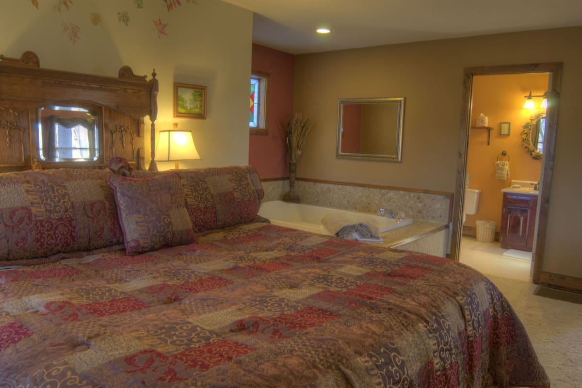 Une chambre à coucher avec un grand lit, une fenêtre, un miroir et une salle de bain visible à travers une porte ouverte.