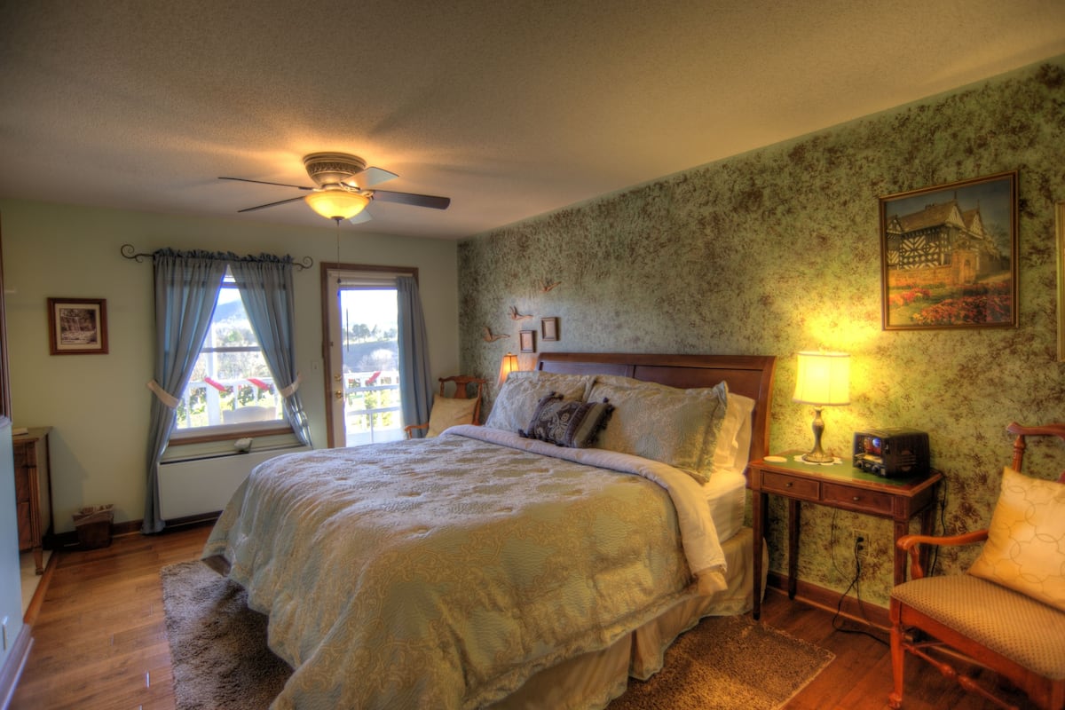 Une chambre avec un grand lit, un ventilateur de plafond, une fenêtre avec des rideaux, une table de chevet avec une lampe et une chaise.