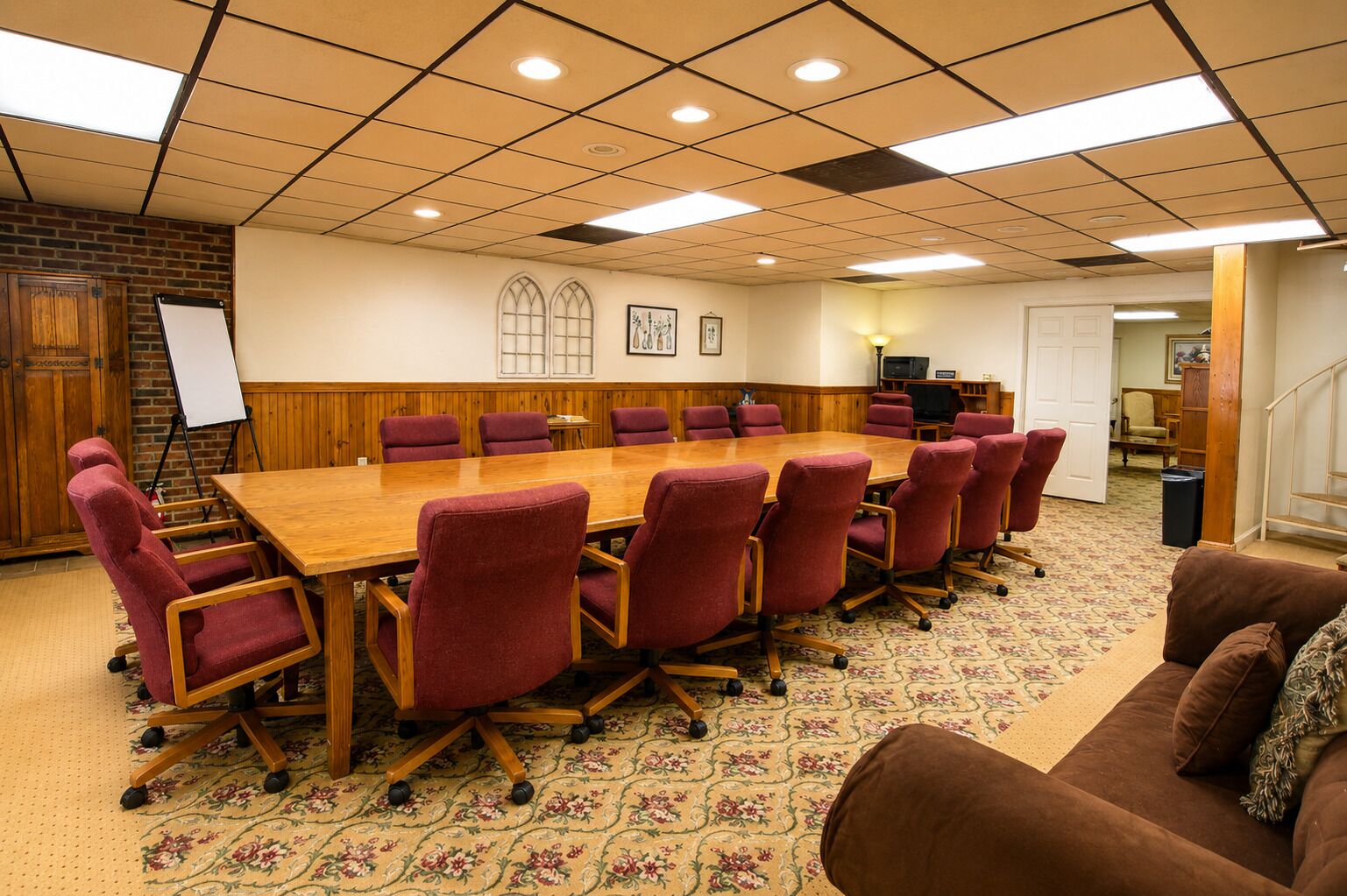 A conference room with a large wooden table, red chairs, a whiteboard, and a carpeted floor.
