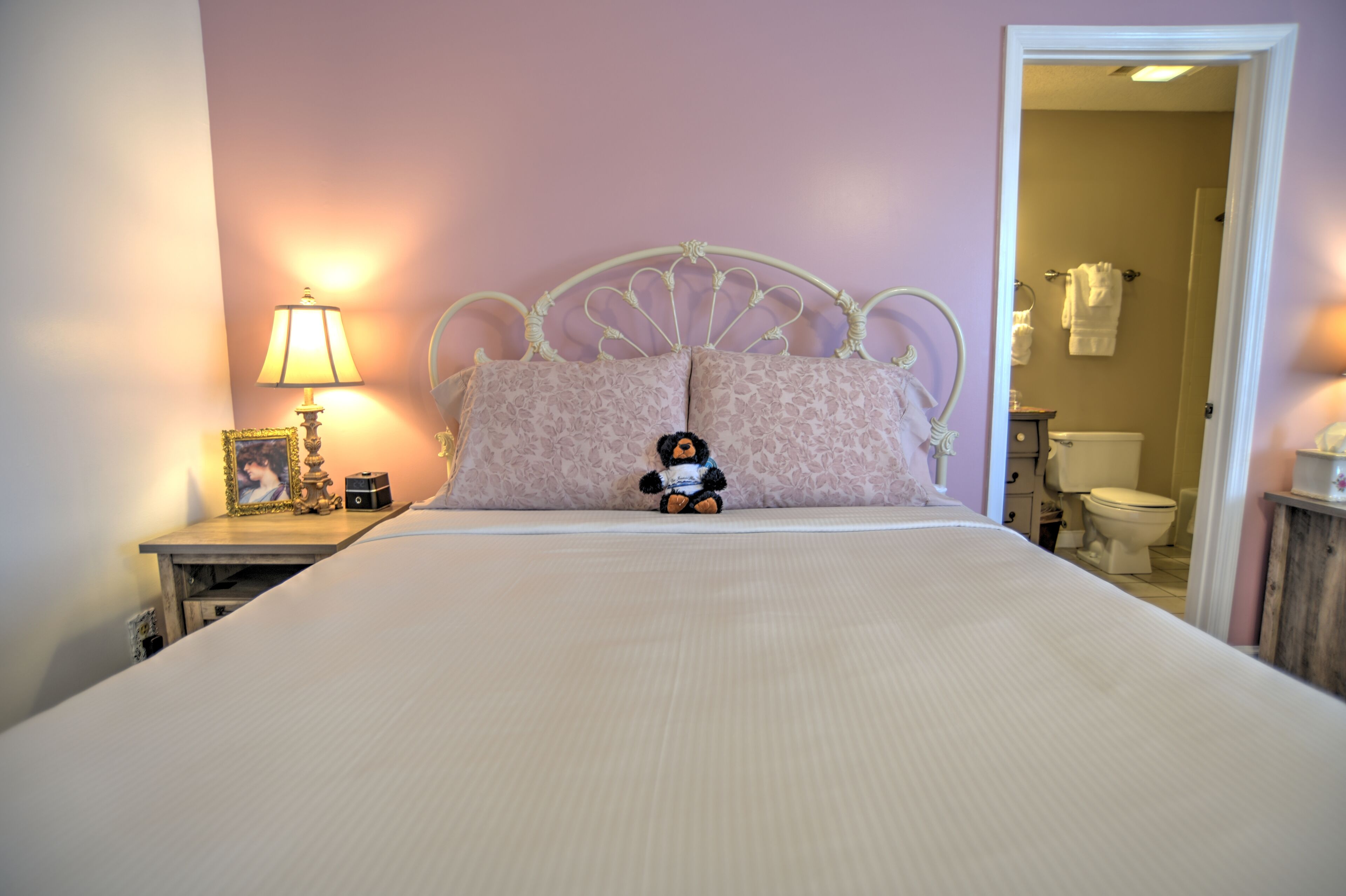 Een net opgemaakt bed met een hoofdbord, een bedlampje en een pluche knuffel.