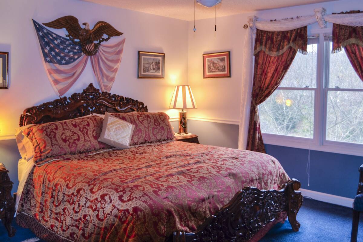 Une chambre à coucher avec un grand lit, un ventilateur de plafond, une fenêtre avec des rideaux et un drapeau américain accroché au mur.