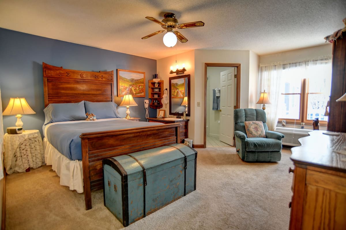 Une chambre avec un grand lit, une tête de lit en bois, un ventilateur de plafond, une commode, une chaise et une fenêtre avec des rideaux.