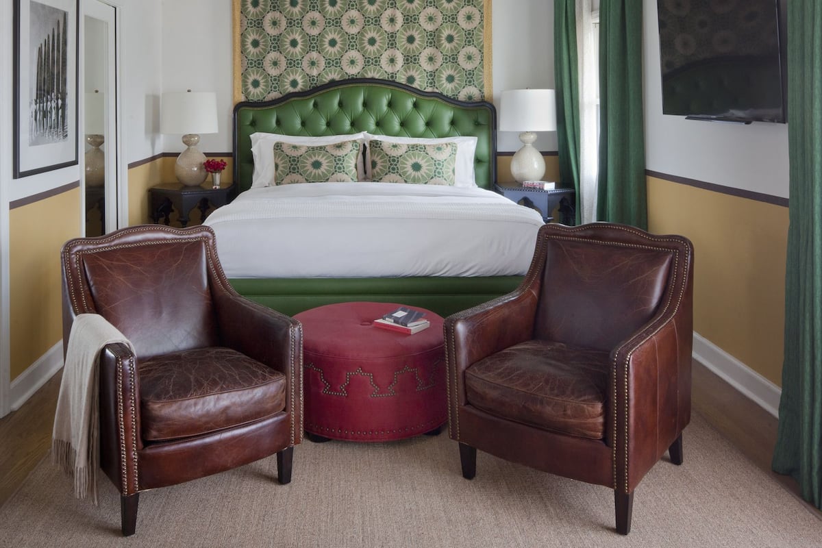 Quarto de hotel com cama estofada em verde, duas poltronas de couro, um banquinho vermelho, televisão fixada na parede e cabeceira com padrão.