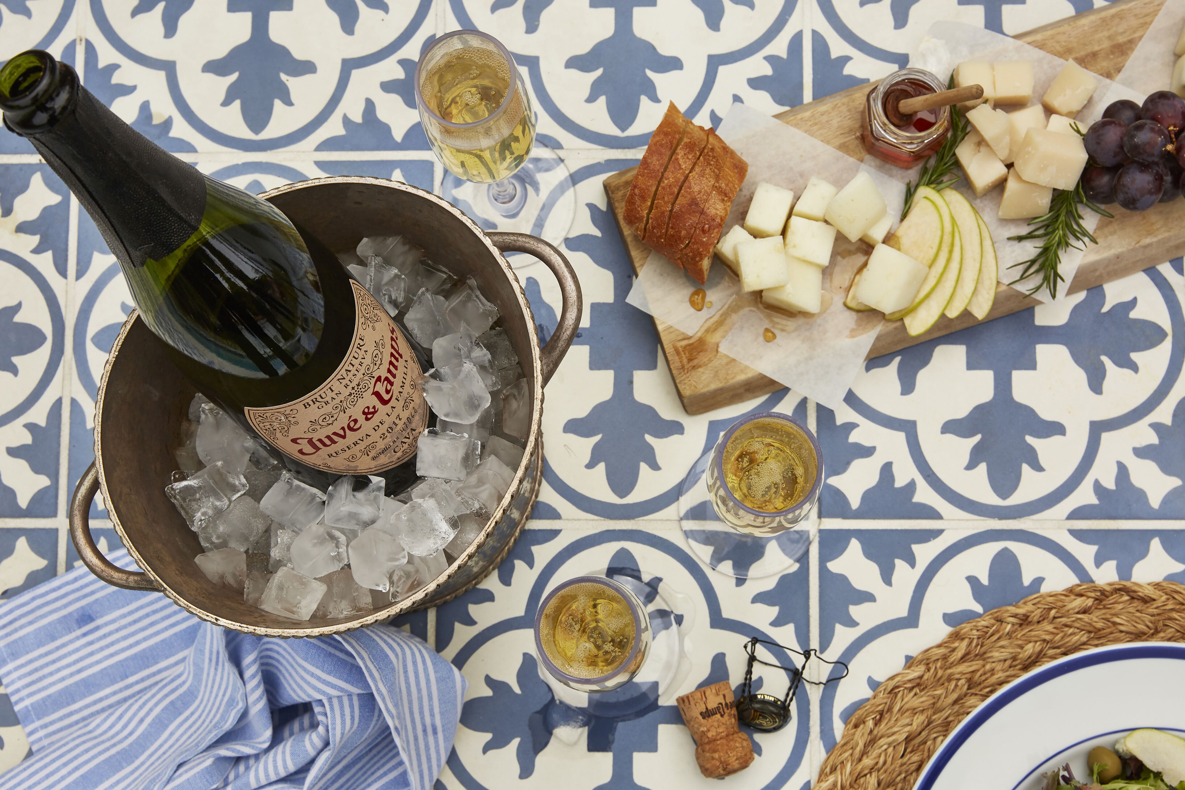 Eine Flasche Champagner, Eiswürfel, Käse, Trauben und eine Weinflasche auf einer gemusterten Tischdecke.