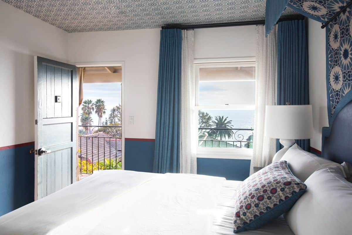 Quarto de hotel com uma cama, janela com cortinas, vista para palmeiras e uma varanda.