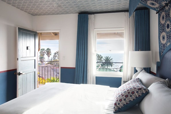Habitación de hotel con cama, ventana con cortinas, vistas a palmeras y un balcón.