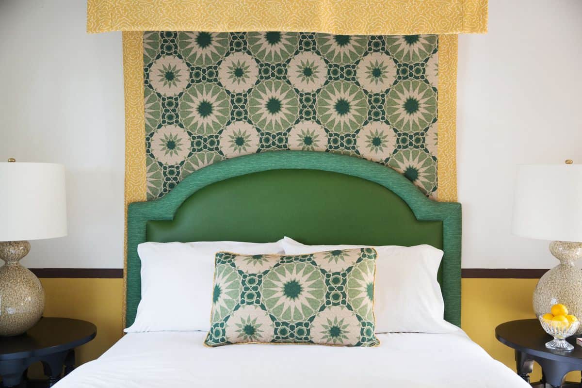 Quarto de hotel com cabeceira verde, tapeçaria de parede estampada, colcha branca e duas mesas de cabeceira com luminárias e vasos decorativos.