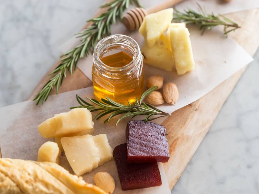 A wooden board with various cheeses, a jar of honey, almonds, and rosemary sprigs.