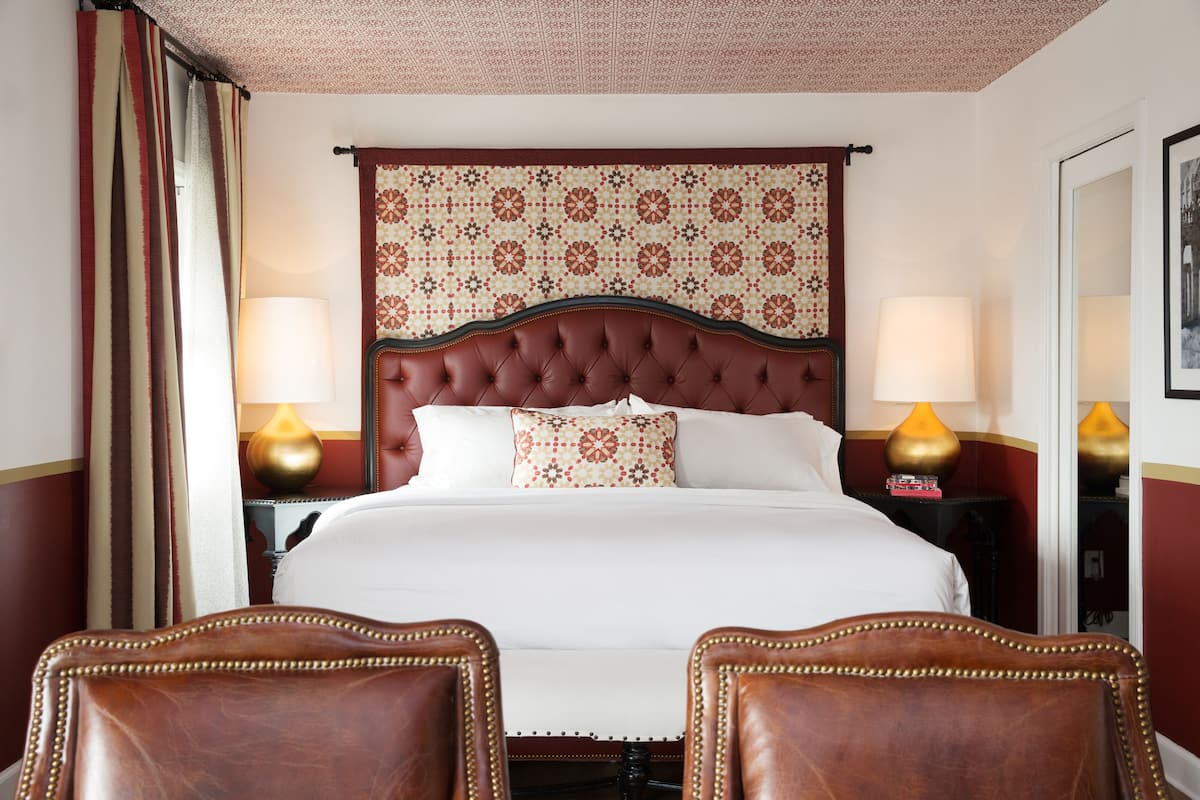 Quarto de hotel com uma cama grande, duas luminárias de cabeceira, um painel de cabeceira estampado e duas poltronas de couro.