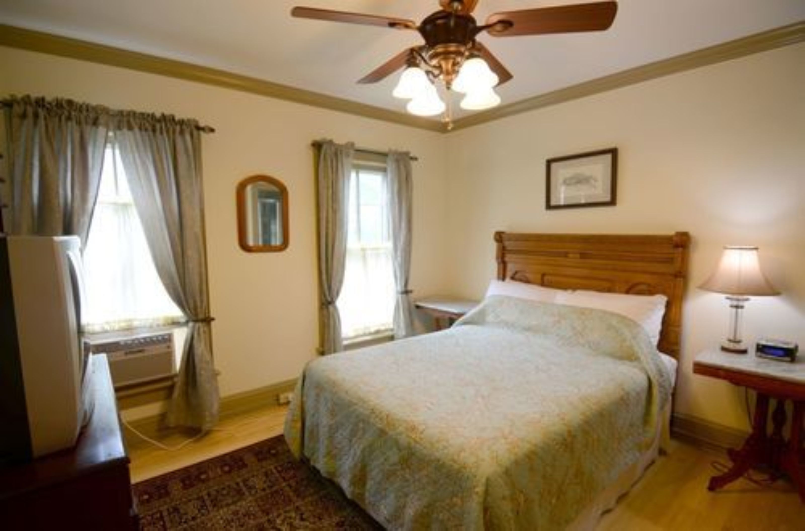 Une chambre à coucher avec un lit, un ventilateur de plafond, une table de chevet avec une lampe et une fenêtre avec des rideaux.