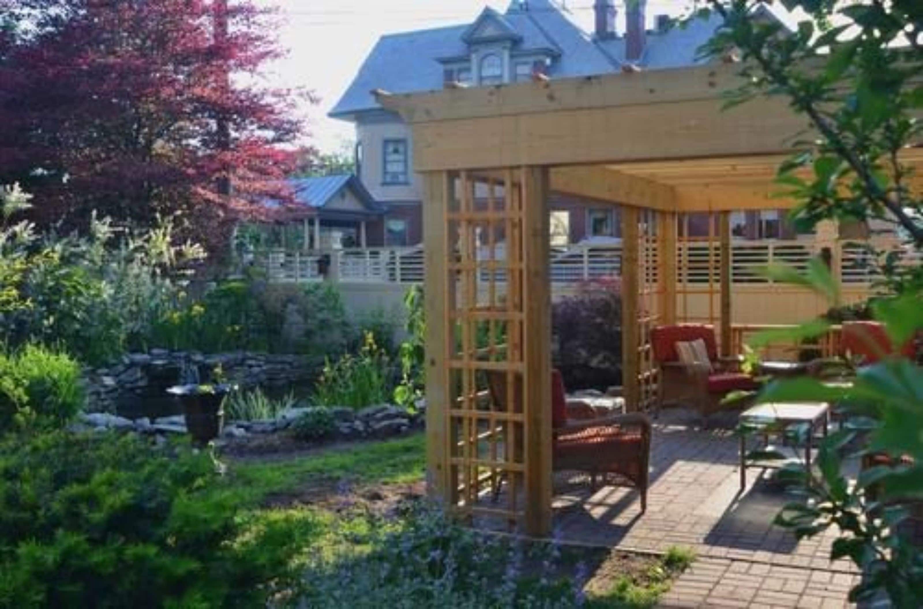 A covered outdoor seating area with a pergola structure, surrounded by lush greenery and a stone feature.