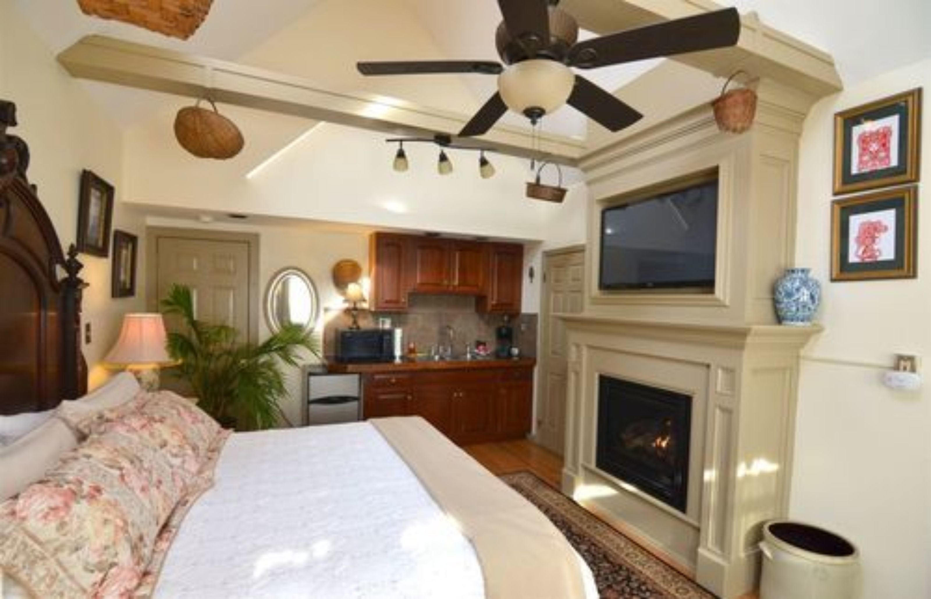 A bedroom with a bed, a ceiling fan, a fireplace, and a kitchen in the background.