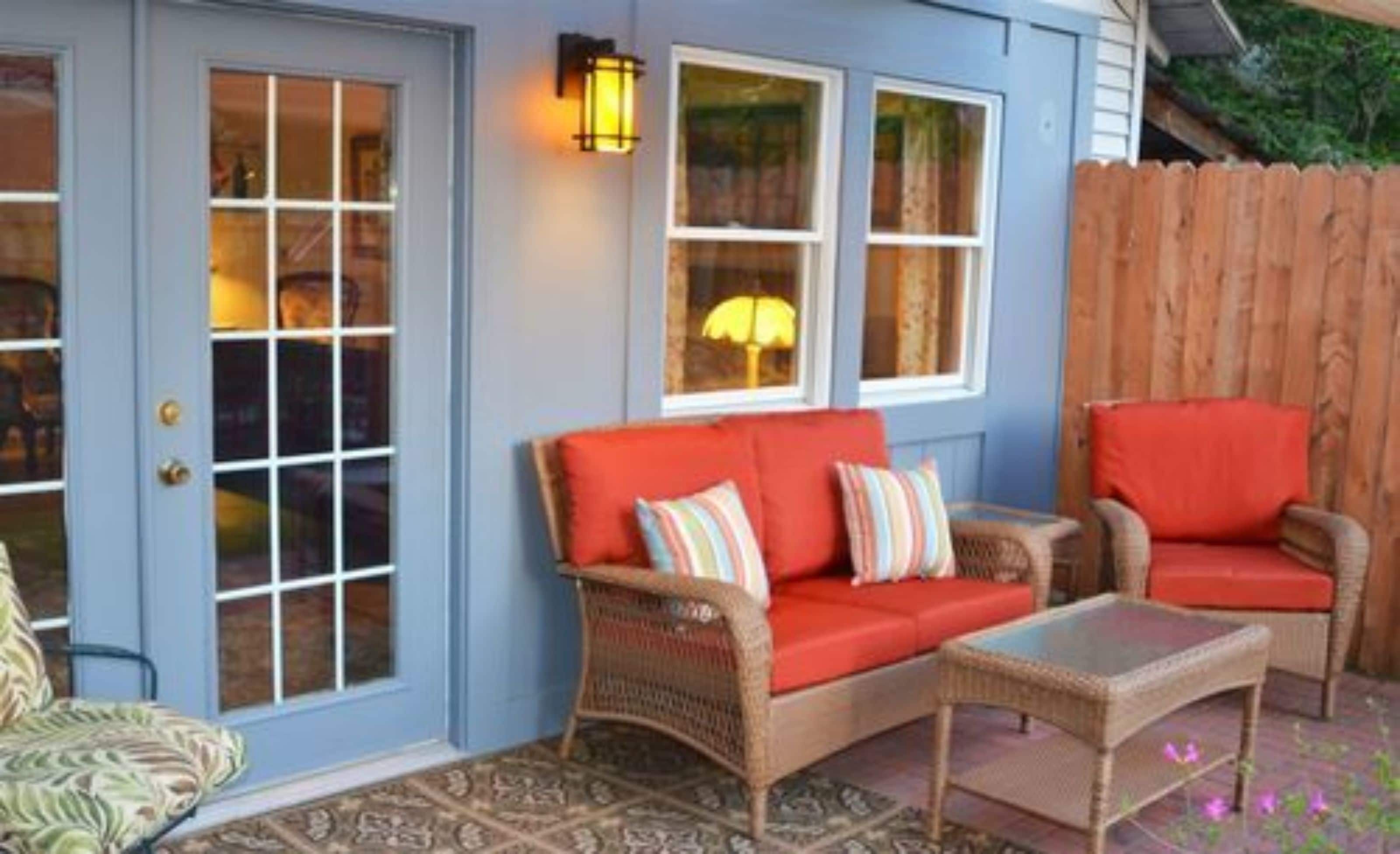 A cozy outdoor seating area with wicker furniture and a red sofa, a wooden fence, and a blue building with windows.
