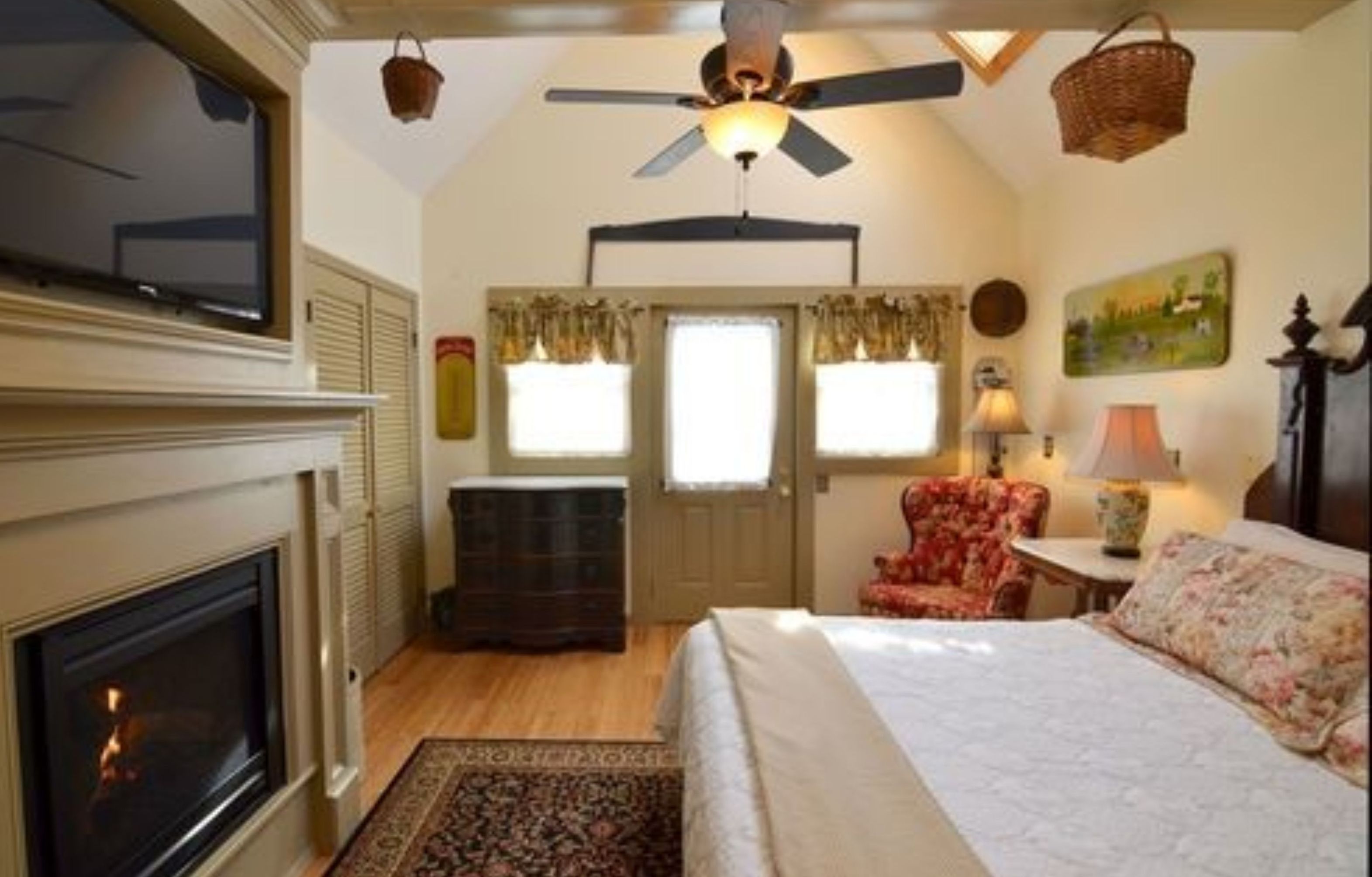 A bedroom with a fireplace, a ceiling fan, a bed, a chair, and a window with curtains.