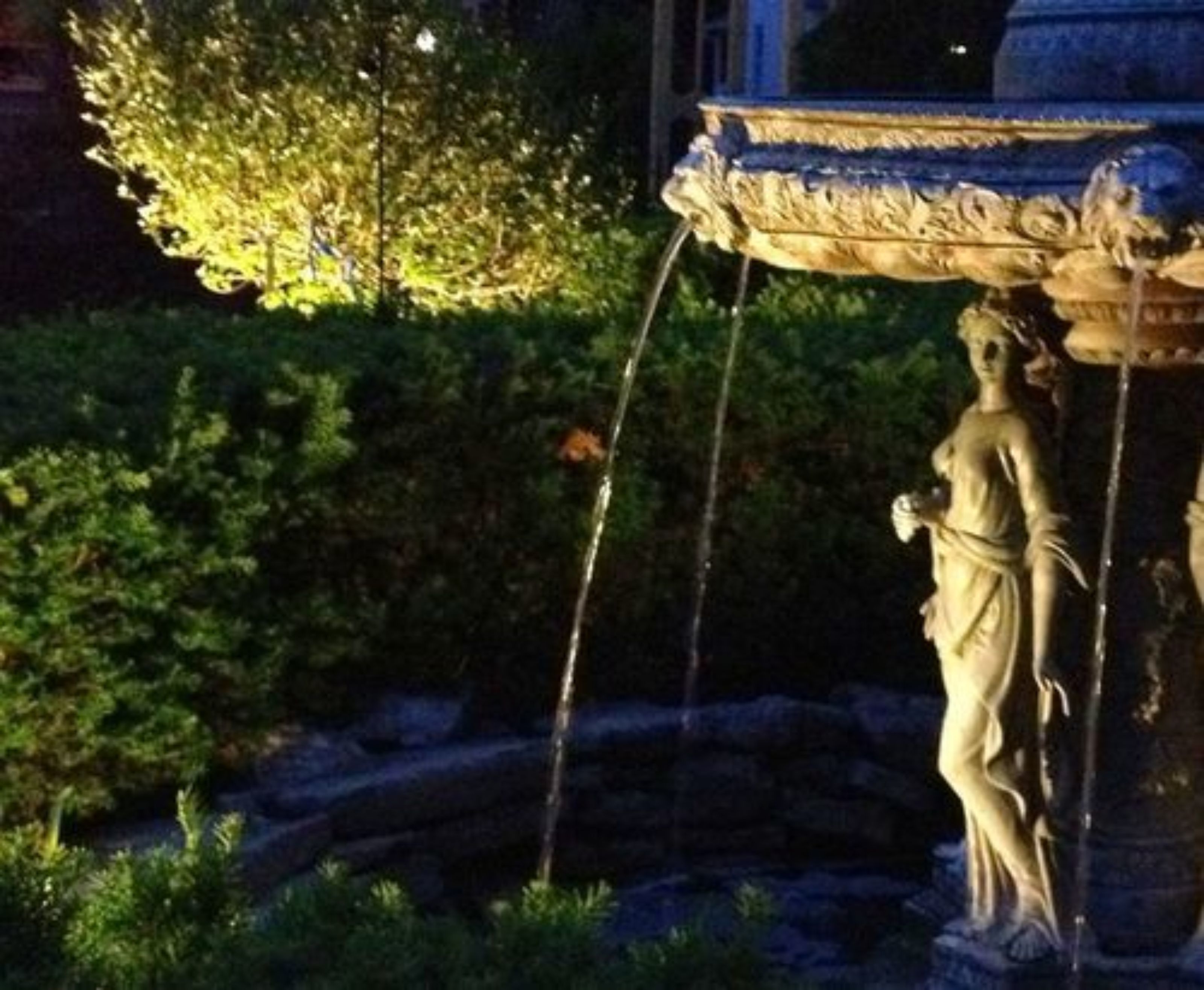 Uma estátua de mulher segurando um bastão, em cima de um pedestal de pedra com uma fonte acima.
