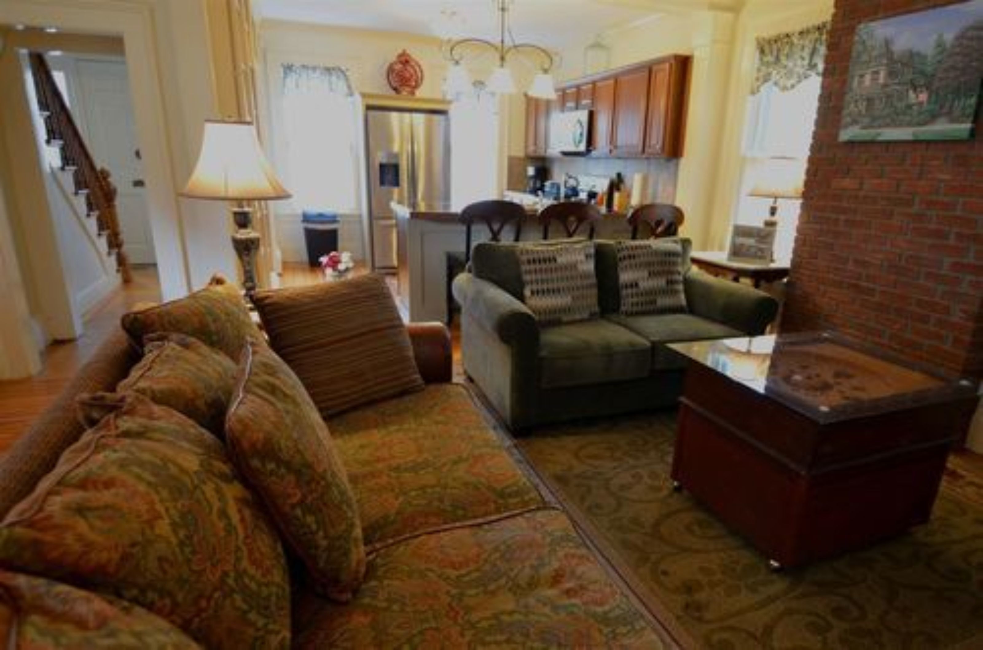 A living room with a sofa, a coffee table, and a fireplace. There is a kitchen in the background with wooden cabinets and a refrigerator.