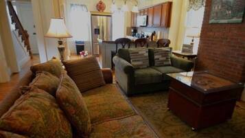 A living room with a sofa, a coffee table, and a fireplace. There is a kitchen in the background with wooden cabinets and a refrigerator.