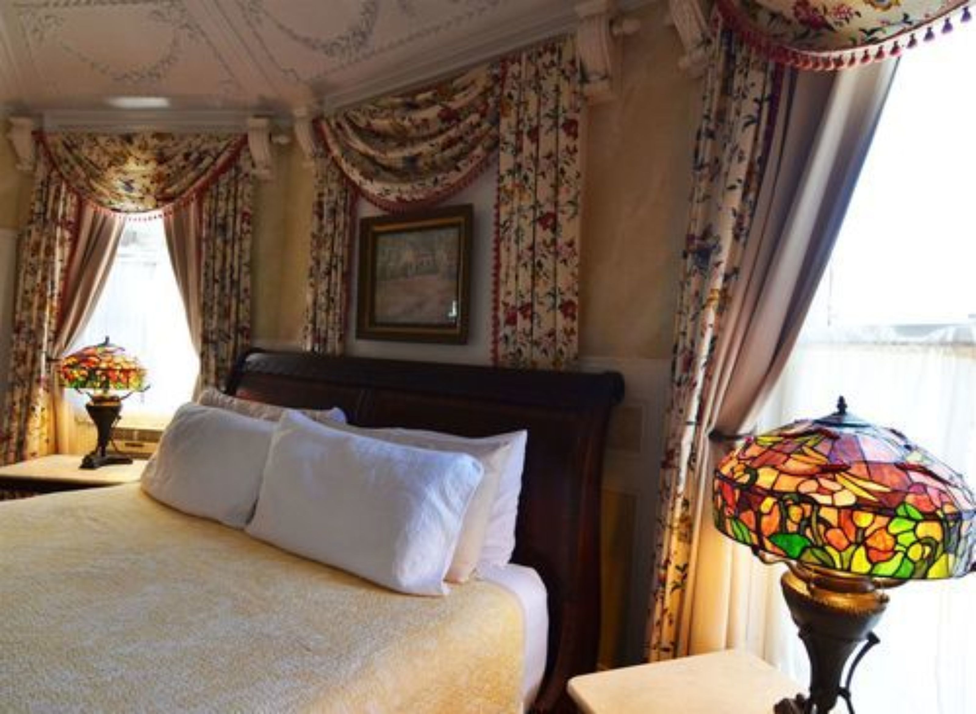 A neatly made bed with white linens in a room with floral curtains and a stained glass lamp.