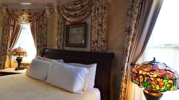 A neatly made bed with white linens in a room with floral curtains and a stained glass lamp.