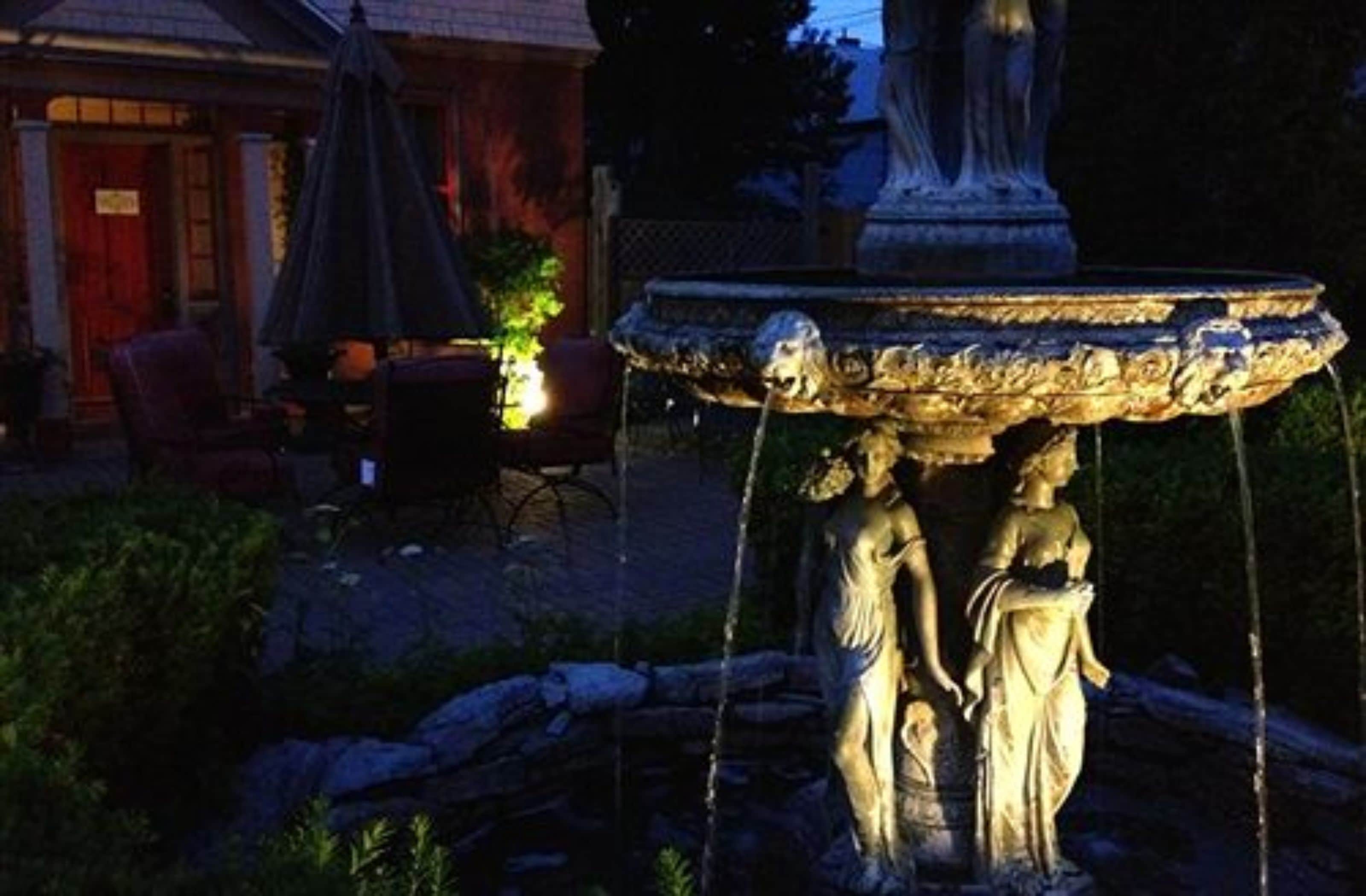 Una fuente de piedra con estatuas, un edificio rojo y mesas al aire libre al fondo.