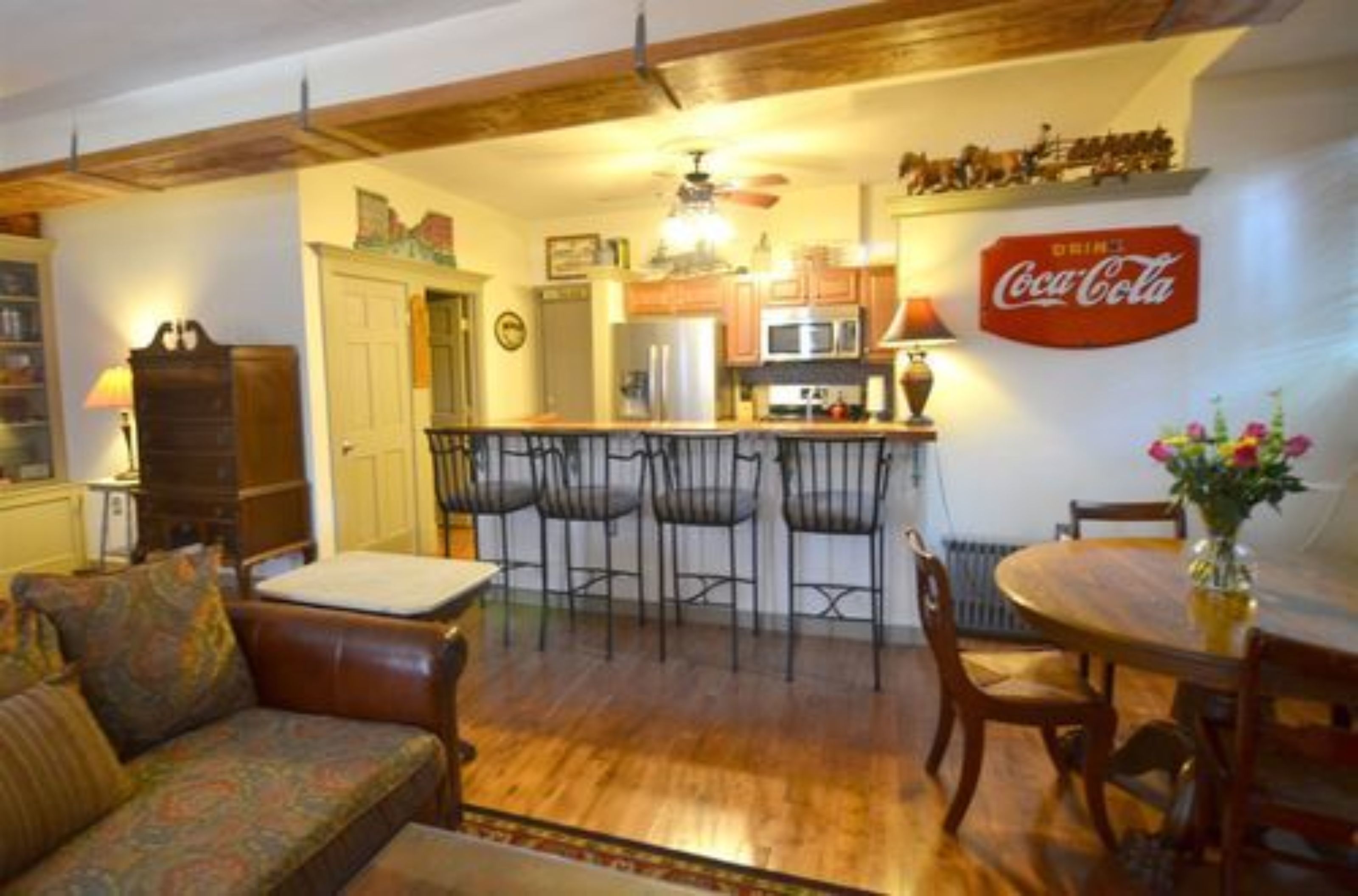 A cozy living room with a sofa, a dining table with chairs, a kitchen area with a refrigerator, microwave, and a Coca-Cola sign.