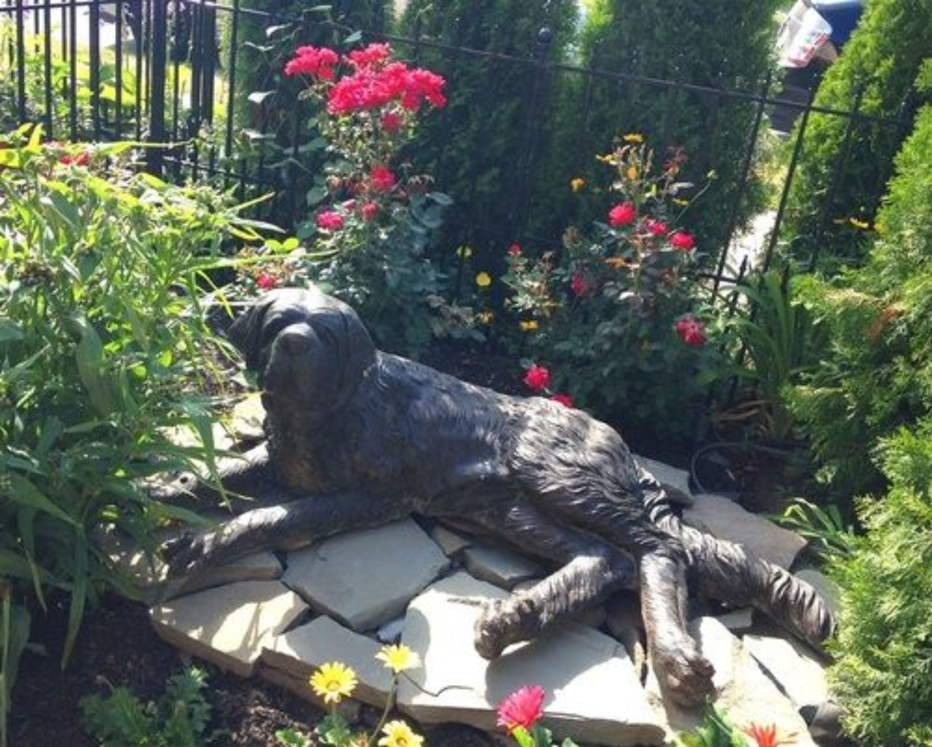 Una estatua de jardín de un perro acostado sobre una losa de piedra, rodeado de flores y follaje.