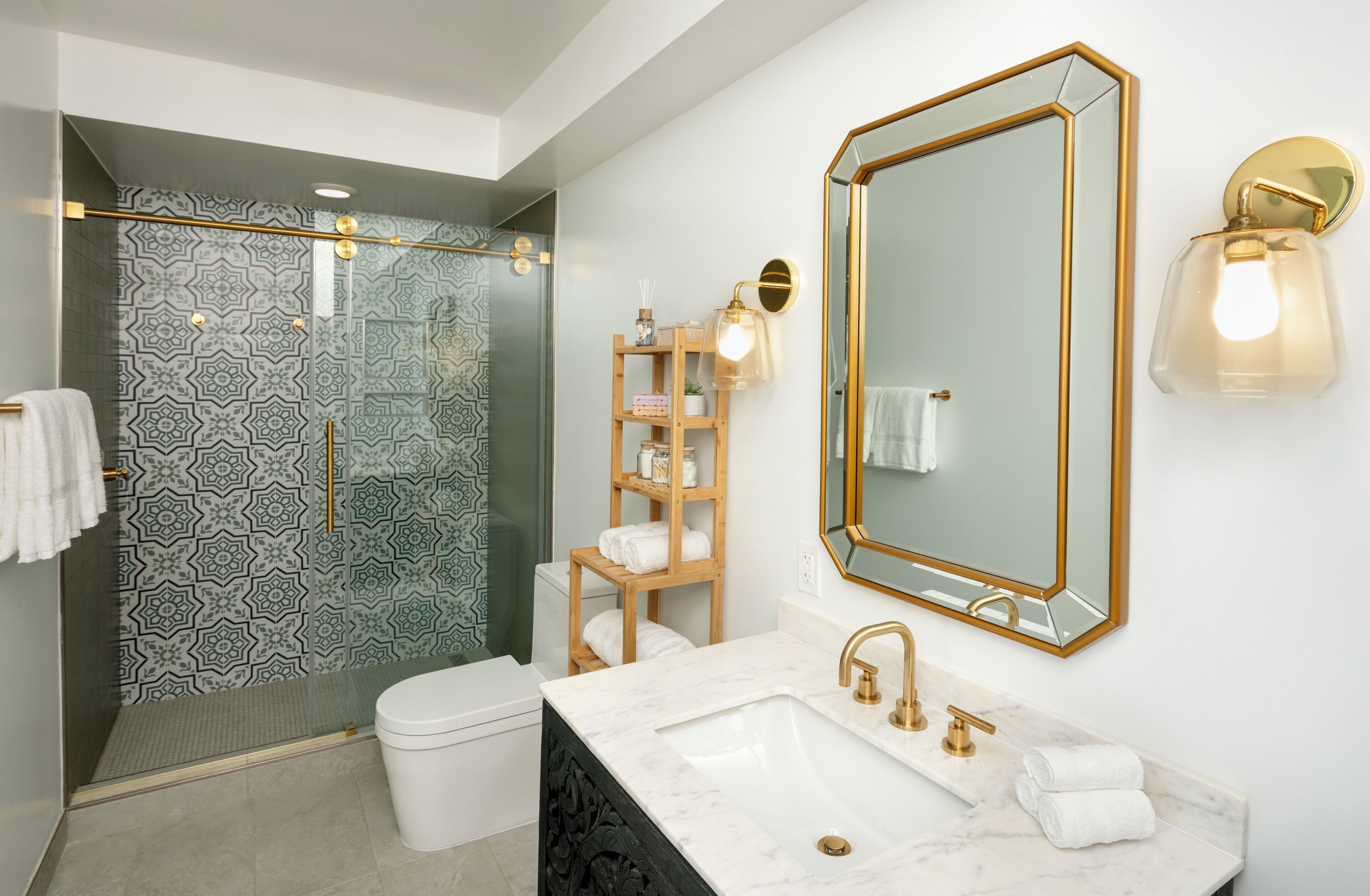 A modern bathroom with a glass shower enclosure, a white toilet, a marble sink with gold fixtures, and a large mirror with a gold frame.