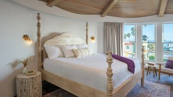 A four-poster bed with a white bedspread and a purple throw blanket. There is a round woven side table with a telephone on it. A window with curtains is visible, and a chair is placed near the window.