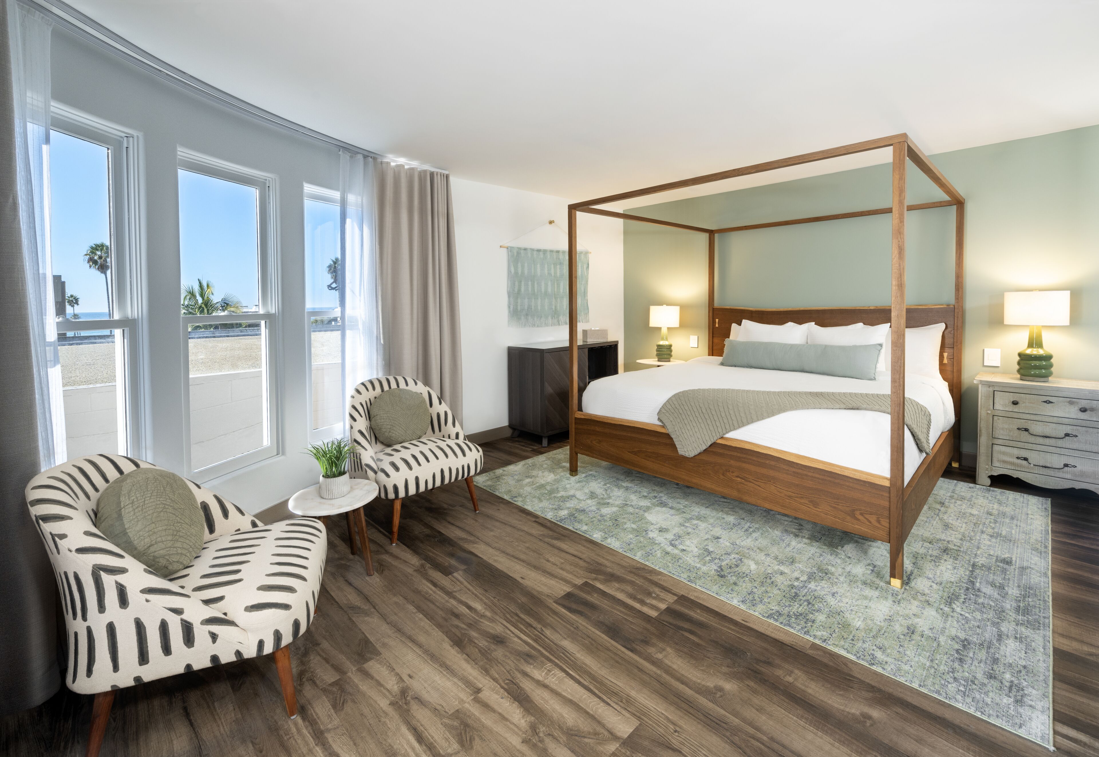 A spacious bedroom with a four-poster bed, a sitting area with a chair and a small table, and a view of the beach through large windows.