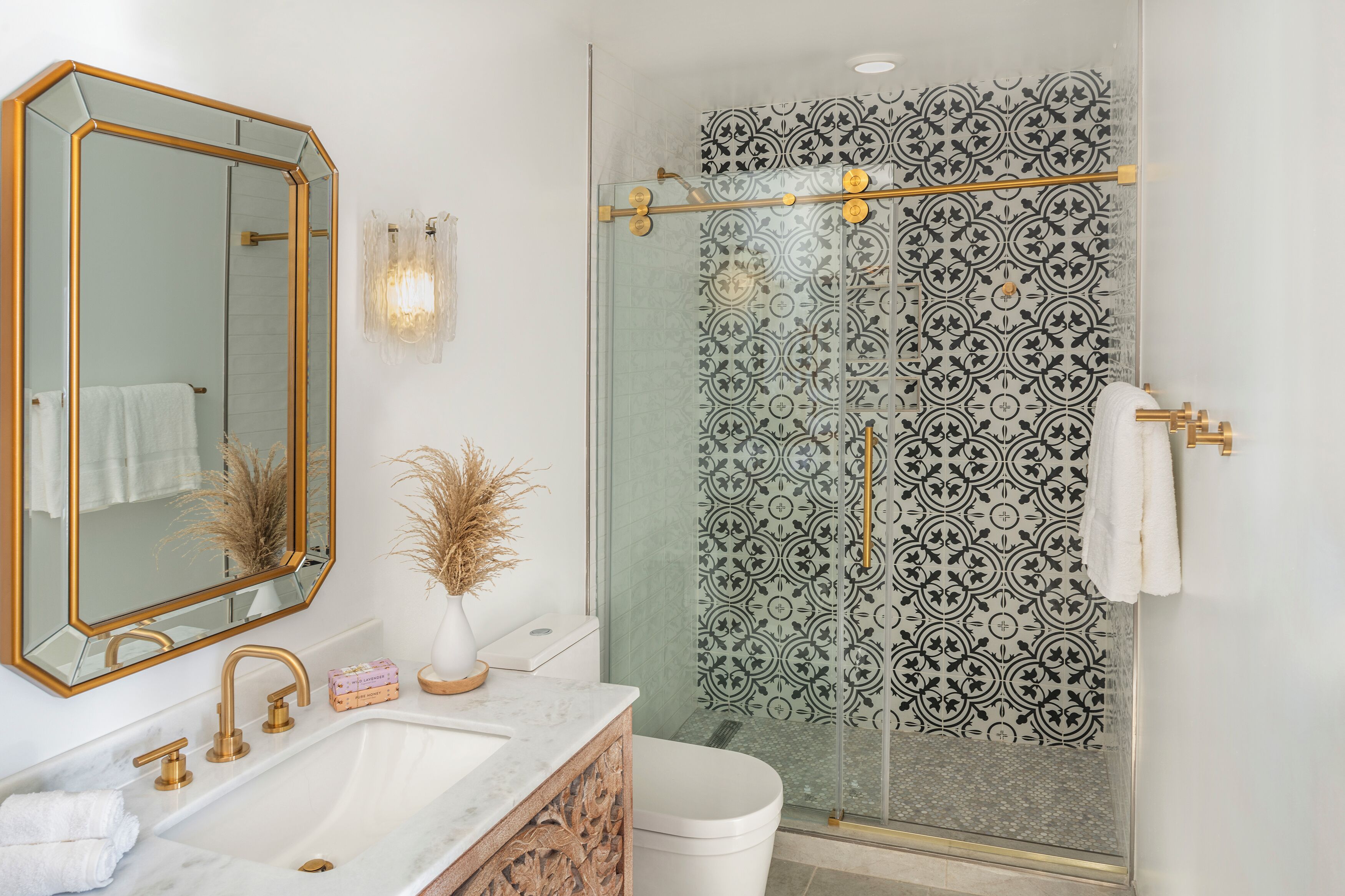 A bathroom with a white sink, gold faucets, a large mirror, and a shower with patterned tiles.