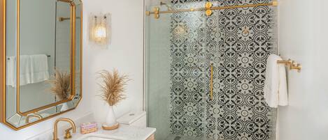 Een badkamer met een wit wastafel, gouden kraanwerk, een grote spiegel en een douche met betegelde wanden.