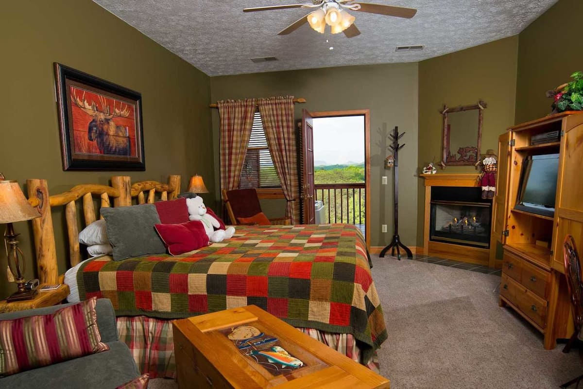 A cozy room with a bed, a wooden desk, a fireplace, and a balcony with a view.