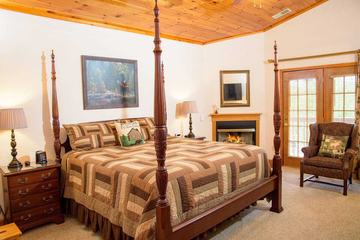 A four-poster bed with a quilt, a nightstand with drawers, a chair with a cushion, a painting on the wall, and a fireplace.