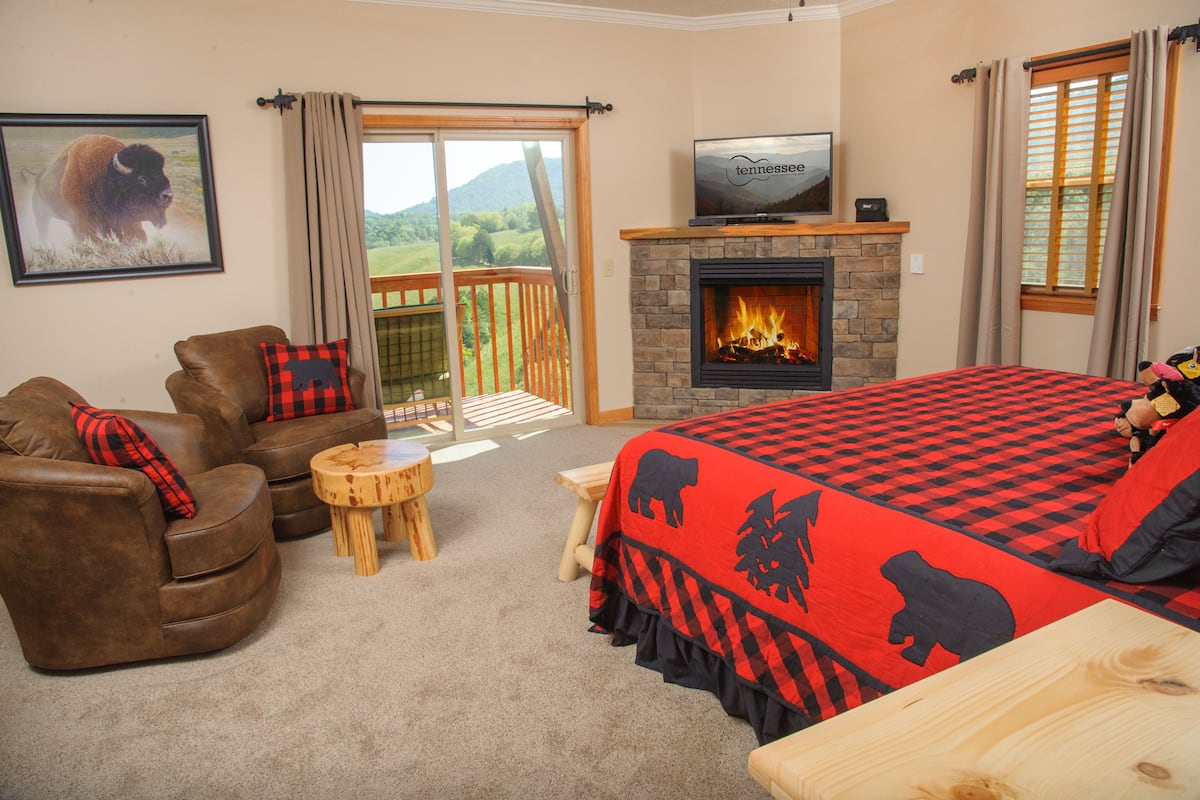A cozy room with a fireplace, a large bed with a checkered bedspread, a sofa with a buffalo pillow, and a television on a mantel.
